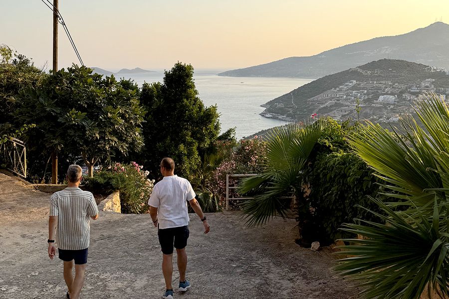 Zwei Männer gehen einen von Pflanzen umgebenen Hügel hinunter zum Meer in Kalkan, und fangen den Charme eines türkischen September-Urlaubs an der Türkisküste ein.