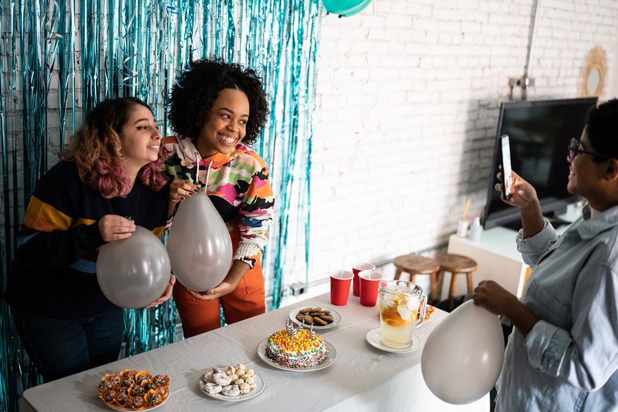 Zwei Mädchen posieren vor einem glitzernden Foto-Hintergrund, halten Luftballons neben einem Snacktisch voller Partyessen und einer Geburtstagstorte, und veranschaulichen posterXXLs Tipps zum Fotografieren von Partydeko und Essensarrangements.