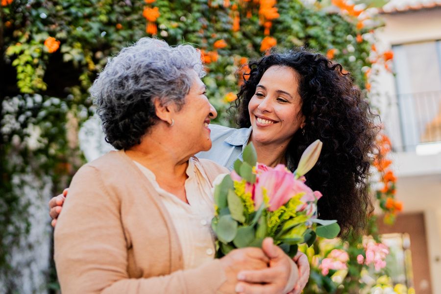 Eine junge Frau lächelt ihre Mutter an, die Blumen in den Händen hält, und veranschaulicht posterXXLs Ideen dafür, was man in eine Hochzeitstagskarte für die Eltern schreiben kann.