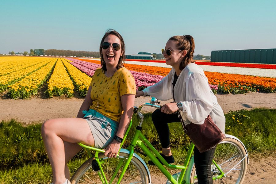 Zwei Frauen fahren gemeinsam Fahrrad, lachen und lächeln in einem blühenden Feld, und veranschaulichen posterXXLs Ideen dafür, was man in eine Hochzeitstagskarte für Freunde schreiben kann.