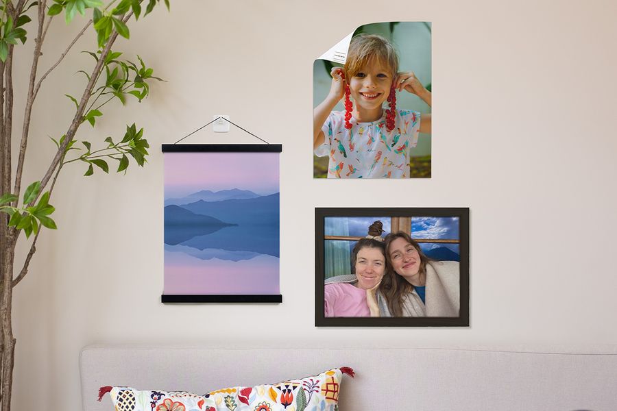 Verschiedene Aufhängeoptionen für Wandkunst von posterXXL werden in einem Zuhause gezeigt, darunter ein personalisiertes Poster eines jungen Jungen, befestigt mit Klebestreifen, ein personalisiertes Landschaftsposter, aufgehängt mit einem Holzbügel und Klebehaken, sowie ein gerahmtes Bild von zwei lächelnden Mädchen.