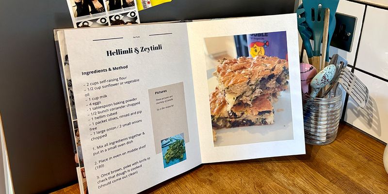 Ein aufgeschlagenes persönliches Kochbuch liegt in einer Küche, mit einem Glas voller Löffel und Küchenutensilien im Hintergrund, und stellt posterXXLs Ratgeber zum eigenen Kochbuch erstellen vor.