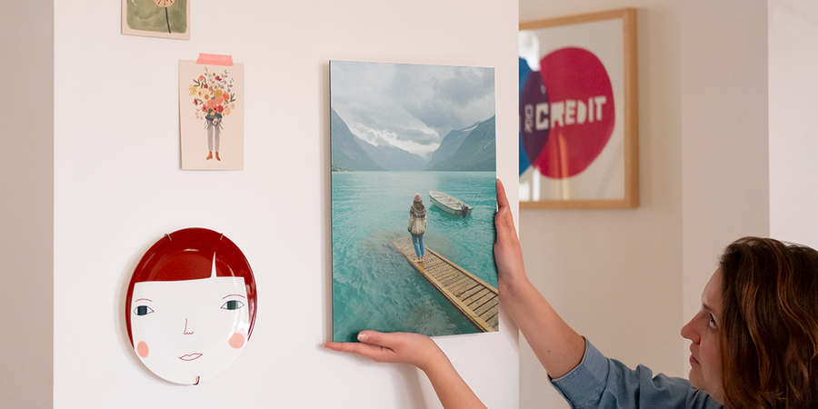Eine Frau hängt personalisierte Wandkunst an ihre Wand, neben einem dekorativen Teller und einigen Fotoabzügen, und stellt posterXXLs Schritt-für-Schritt-Anleitung zum Aufhängen von Wandkunst mit Aufhänge-Sets vor.