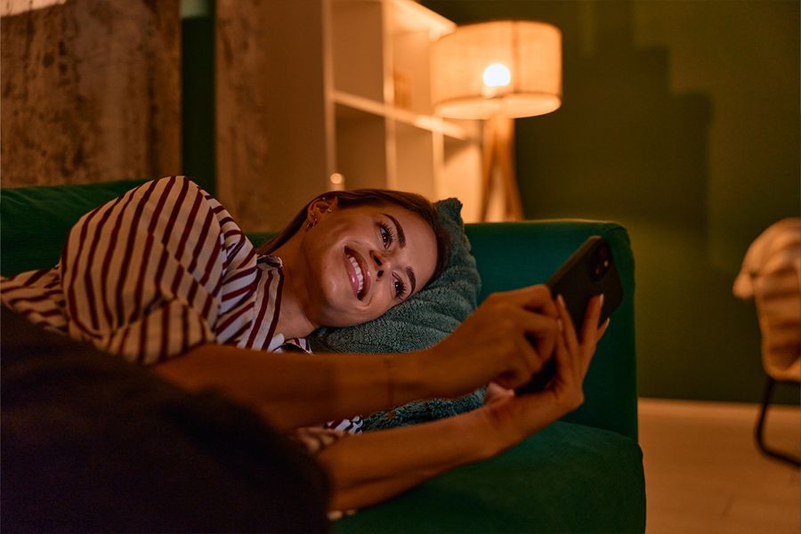Eine Frau liegt auf einem dunkelgrünen Sofa und schaut in einem Wohnzimmer auf ihr Handy, beleuchtet von einer Lampe für gemütliches Licht, und veranschaulicht posterXXLs Tipps für Hygge-Beleuchtung zu Hause.