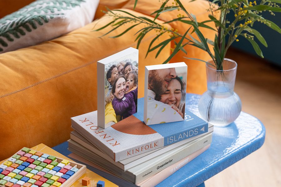 Zwei personalisierte Fotoblöcke liegen auf einer Auswahl an Bildbänden auf einem blauen Beistelltisch neben einem orangefarbenen Samtsofa und veranschaulichen, wie posterXXLs Fotoblöcke einem Hygge-Zuhause eine persönliche Note verleihen.