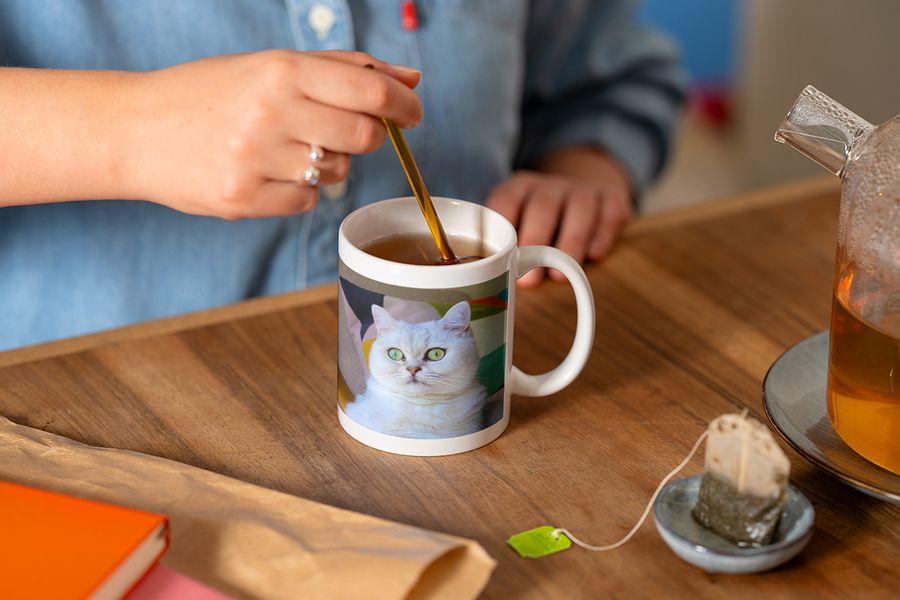 Jemand rührt mit einem Löffel in einer personalisierten Fototasse mit dem Foto einer weißen Katze, während er eine Tasse Tee zubereitet, und veranschaulicht posterXXLs Tipp, eine Hygge-Leseecke mit einer personalisierten Tasse abzurunden.