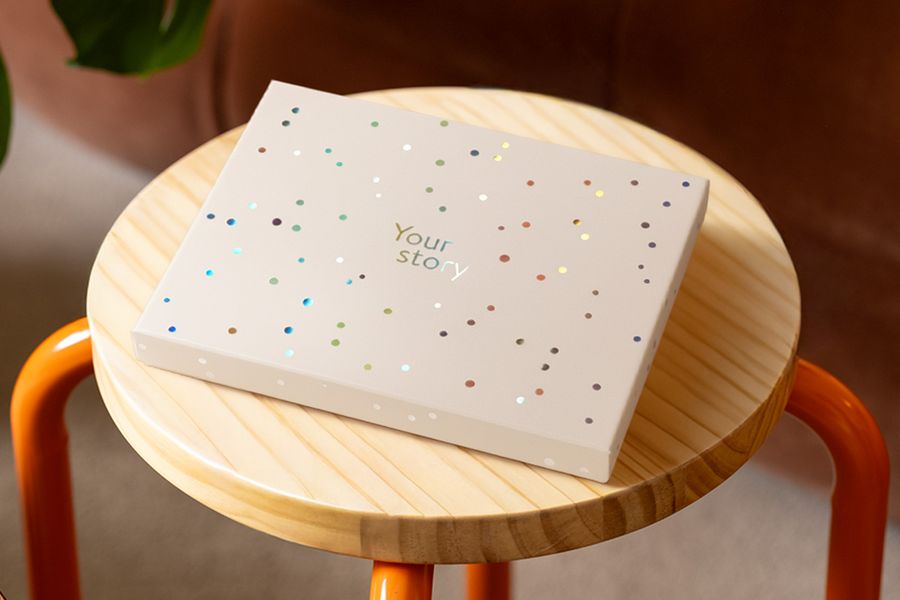 Eine posterXXL Präsentationsbox steht auf einem Holzhocker, verziert mit silberfarbenen holografischen Kreisen und dem Text 'Your Story', und veranschaulicht das matte, elfenbeinfarbene Design mit einem Hauch holografischem Glanz.