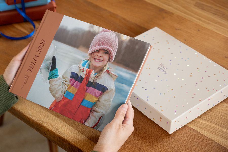 Jemand hält ein Fotobuch mit dem Titel 'Julia', das ein lächelndes Mädchen auf dem Cover zeigt, mit einer Präsentations-Geschenkbox direkt dahinter auf dem Tisch, und veranschaulicht, warum eine Fotobuchbox eine Kreation besonders macht.