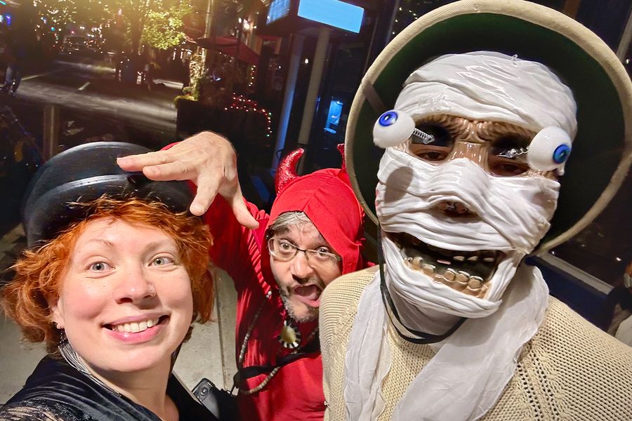 Drei Erwachsene in Halloween-Kostümen auf der Straße, einer mit einer Skelettmaske mit hervorquellenden Augen, einer als Teufel verkleidet und eine Frau mit schwarzem Hut, und veranschaulichen posterXXLs Halloween-Sprüche für Kostüme.