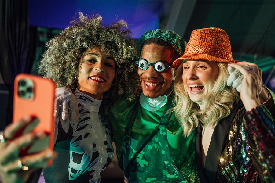 Drei Erwachsene posieren für ein Selfie auf einer Halloween-Party, eine Frau im Skelettkostüm, ein Mann als gruseliger Chirurg mit lustiger Brille und eine Frau mit Pailletten-Hut, und veranschaulichen posterXXLs Halloween-Sprüche für Gruppen.