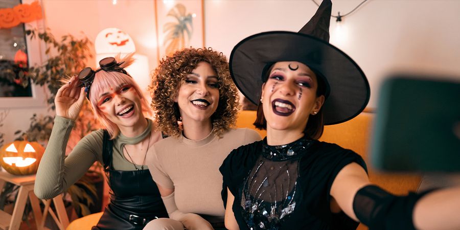 Drei Frauen in Kostümen auf einer Halloween-Party, eine Hexe, ein Tier und eine Cyberpunk, posieren für ein Selfie mit dem Handy, und stellen posterXXLs 105 Halloween-Sprüche für Instagram vor.