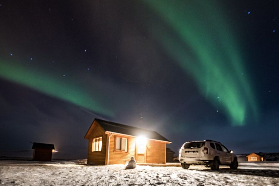 Eine Hütte mit einem hellen Licht, das nach draußen scheint, neben einem Auto in einer sehr verschneiten Landschaft, darüber grüne Polarlichter am Himmel, zur Illustration von posterXXLs Erklärung, was Polarlichter sind und was Aurora borealis verursacht.