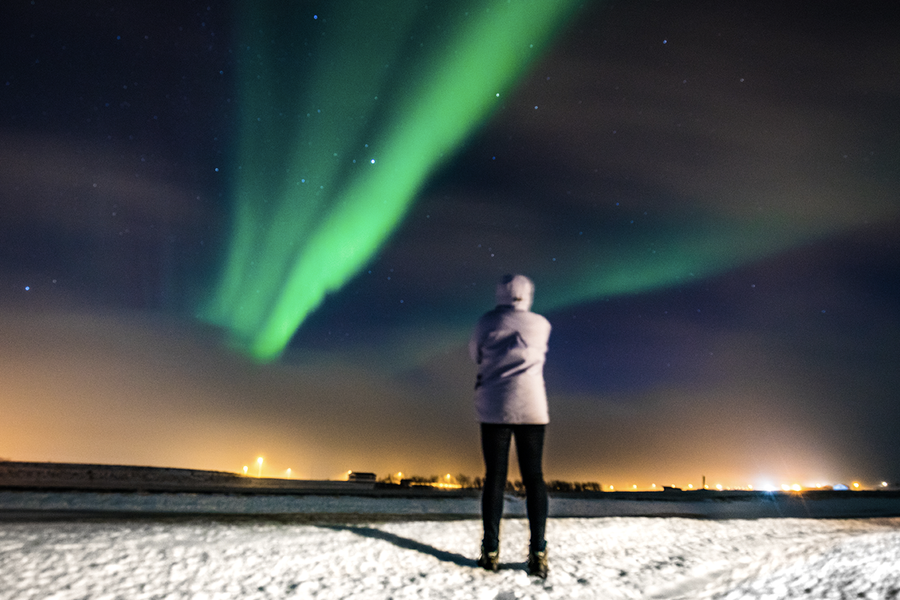 Eine Person steht in einer verschneiten Landschaft und blickt zu den grünen Polarlichtern am Himmel, zur Illustration von posterXXLs Tipps zum Fotografieren der Polarlichter mit dem Handy.