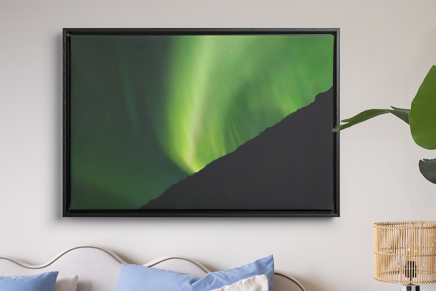 Ein gerahmter Druck von sehr grünen Polarlichtern hängt an der Wand über einem Bett und einer Raffialampe, zur Illustration von posterXXLs Wandkunst, mit der sich Polarlichtfotos in etwas Bleibendes verwandeln lassen.