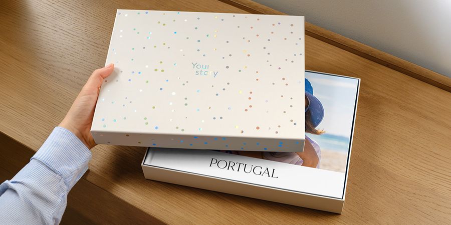 Eine posterXXL Präsentationsbox steht mit leicht geöffnetem Deckel, der den Blick auf ein personalisiertes Reisefotobuch für Portugal im Inneren freigibt, dessen Cover teilweise vom Deckel verdeckt wird, und stellt posterXXLs neue Fotobuch-Geschenkbox vor.