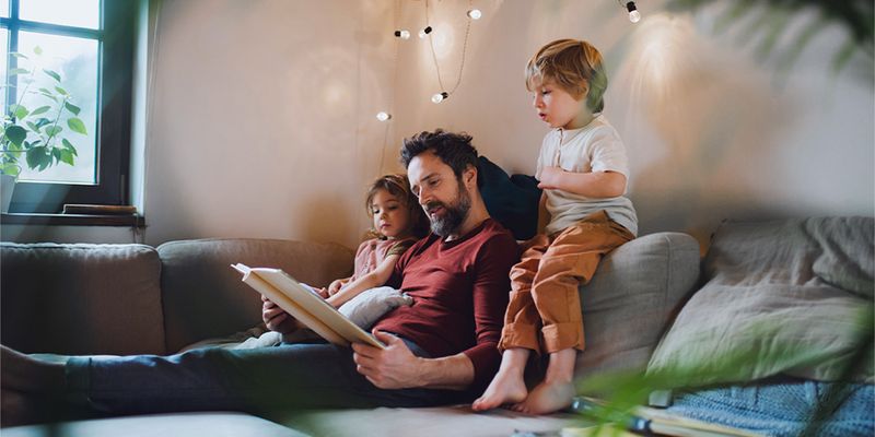 Ein Vater liest einem kleinen Jungen und einem kleinen Mädchen auf dem Sofa ein Buch vor, mit warmen Lichterketten vor ihnen, und stellt posterXXLs Tipps für gemütliche Herbstdeko im Hygge-Stil vor.