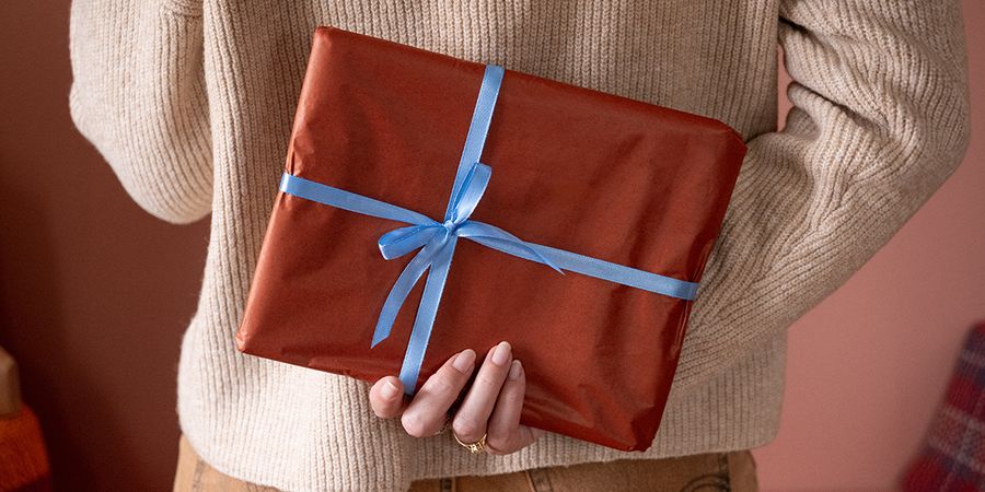Frau, die hinter ihrem Rücken ein in braunes Papier eingepacktes Geschenk mit blauem Band hält, passend zu posterXXLs Checkliste mit Weihnachtsgeschenk-Ideen für ein personalisiertes Fotogeschenk.