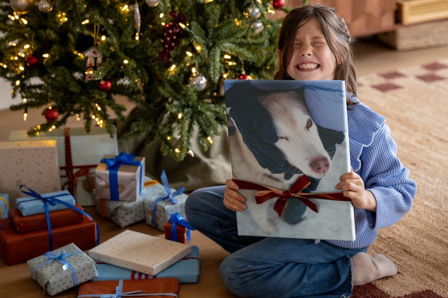 Ein lachendes Mädchen in einem blauen Pullover hält einen Leinwanddruck mit einem Hundefoto hoch, der mit einer roten Schleife verziert ist, vor einem Weihnachtsbaum, als Illustration für posterXXLs Tipps zu Wandkunst.