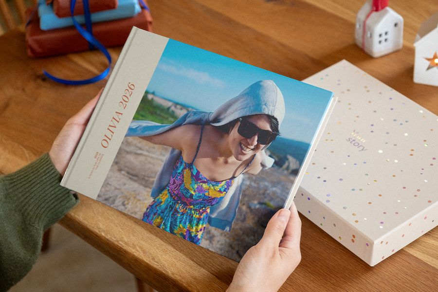 Zwei Hände halten ein Fotobuch mit 2026 auf dem Cover, das eine lächelnde Frau mit Sonnenbrille an einer felsigen Küste zeigt, neben einer Präsentationsbox mit Konfetti-Punkten, als Illustration für posterXXLs personalisierte Fotobücher.
