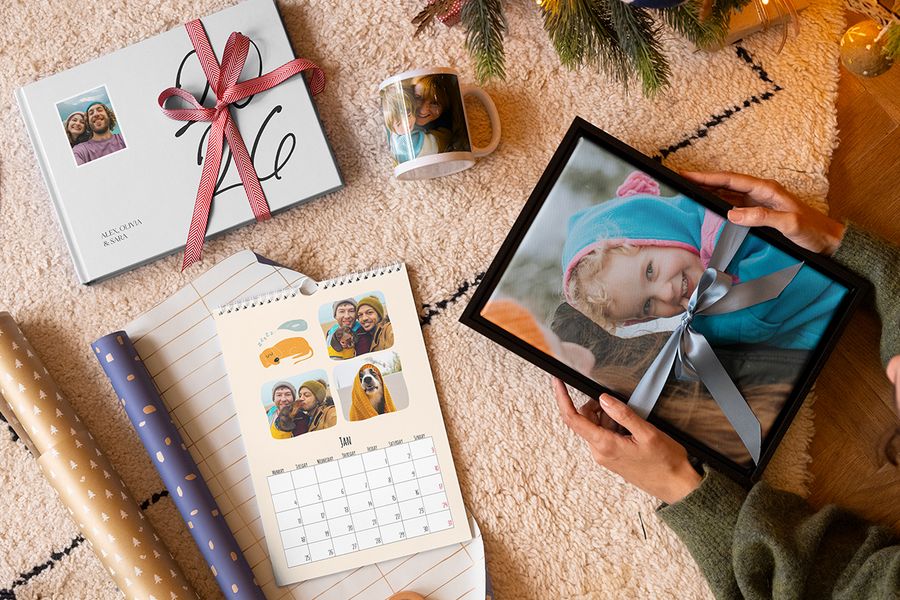 Auswahl personalisierter Geschenke von posterXXL, darunter Wandkunst mit einem jungen Mädchen als Motiv und einer blauen Schleife darum, eine Tasse mit Mutter und Kind, ein personalisierter Fotokalender und ein Fotobuch 2026 mit roter Schleife, neben Geschenkpapier und mit Zweigen eines Weihnachtsbaums im Hintergrund, passend zu posterXXLs Tipps, um die richtigen Erinnerungen in einem personalisierten Weihnachtsgeschenk festzuhalten.