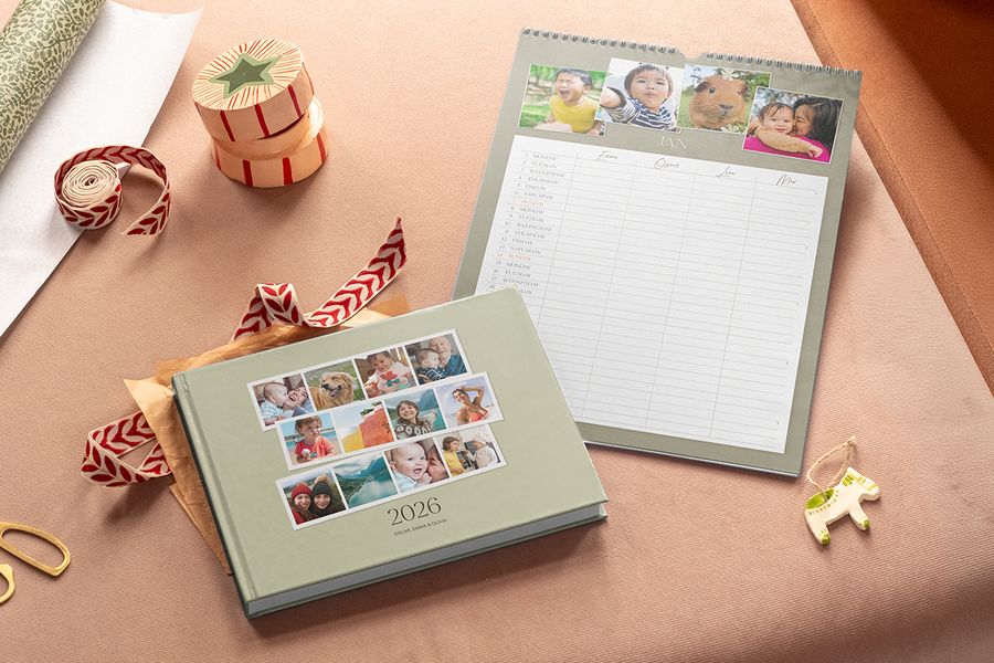 Ein salbeigrünes Fotobuch mit 2026 auf dem Cover und ein Familien-Wandkalender mit Fotos oben und Spalten für jedes Familienmitglied, als Illustration für posterXXLs gemeinsamen Familienkalender zu Black-Friday-Weihnachtsgeschenken.