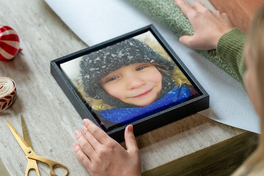 Gerahmte Leinwand mit einem jungen Mädchen in Winterkleidung im Schnee, mit den Händen einer Frau, die bereit sind, sie einzupacken, neben Geschenkpapier, Schere und Band, passend zu posterXXLs Tipps, um ein personalisiertes Fotogeschenk passend zum Stil des Beschenkten auszuwählen.
