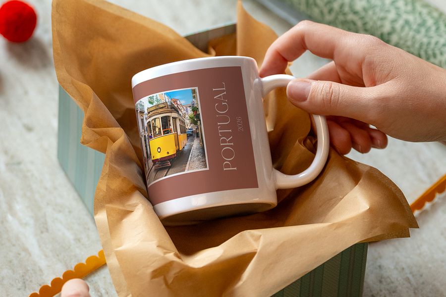 Eine Hand hebt eine Tasse hoch, die in Kraftpapier verpackt und mit einer gelben Straßenbahn und dem Schriftzug „Portugal 2026“ bedruckt ist, als Illustration für posterXXLs personalisierte Tassen mit Hinweis auf ein Erlebnisgeschenk.