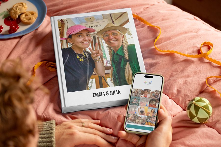 Personalisiertes Fotobuch mit dem Titel “Emma and Julia” mit einer Mutter und ihrer erwachsenen Tochter auf dem Cover, daneben die Hände eines Mannes, der ein Handy hält, um zu zeigen, wie einfach es ist, Fotos auszuwählen und ein Fotobuch von posterXXL zu gestalten, passend zu posterXXLs Tipps, um Fotos von Familie und Freunden für ein personalisiertes Geschenk zu sammeln.
