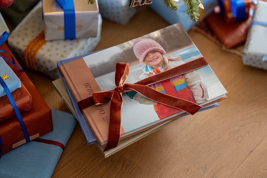 Ein Stapel Fotobücher, mit einem Samtband verschnürt, das oberste Buch mit einem lächelnden Mädchen in einer rosa Strickmütze auf dem Cover und 2026 auf dem Buchrücken, zwischen eingepackten Weihnachtsgeschenken, als Illustration für posterXXLs Fotobuch des Jahres.