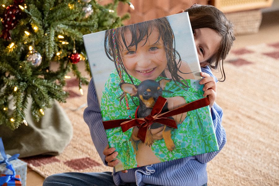 Kleines Mädchen unter dem Weihnachtsbaum, das ein großes Fotobuch mit einem Samtband darum hält, auf dessen Cover ein Foto von ihr mit einem Chihuahua zu sehen ist, passend zu posterXXLs Checkliste mit Weihnachtsgeschenk-Ideen für ein personalisiertes Fotogeschenk.