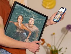 Eine Frau hält ihr Smartphone mit dem posterXXL Editor in der Hand und trägt unter dem Arm Urlaubs-Wanddeko mit zwei Männern, die im Pool Spaß haben – als Inspiration, Urlaubsfotos in schöne Wandbilder zu verwandeln.