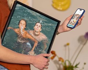 Eine Frau hält posterXXL Wanddeko mit einer Familie, die im Urlaub im Wasser spielt – eine inspirierende Idee, um Urlaubsfotos in schöne Wandbilder zu verwandeln.